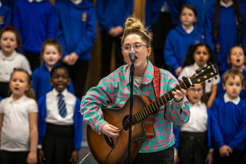 photo shows singer Be Charlotte at a microphone, playing guitar, in front of singing pupils from Our Ladys RC Primary School.