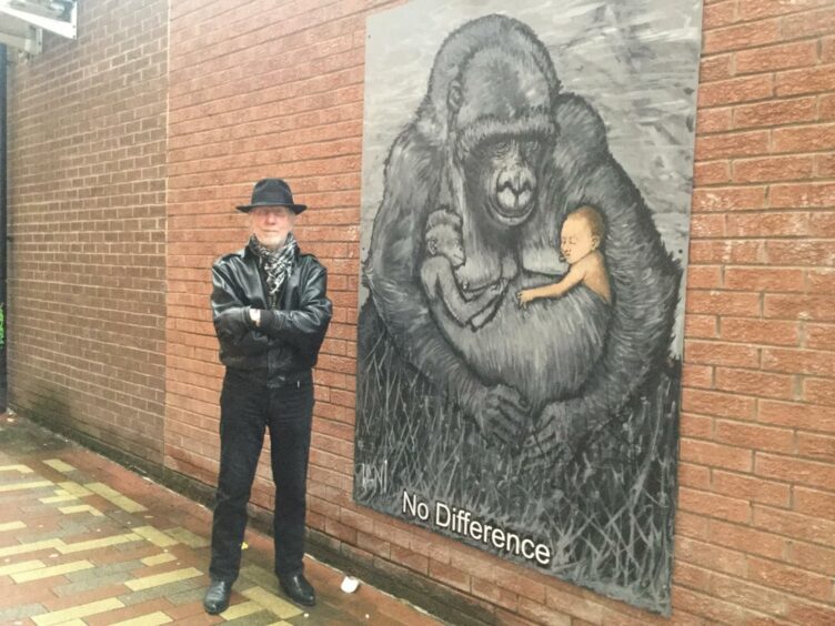 Ian Imrie next to large picture on a brick wall showing gorilla holding baby gorilla and infant child.