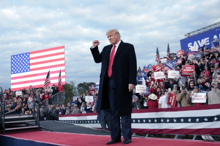Photo shows Donald Trump on stage in front of a crowd holding placards saying 'Save America' and a large American flag.