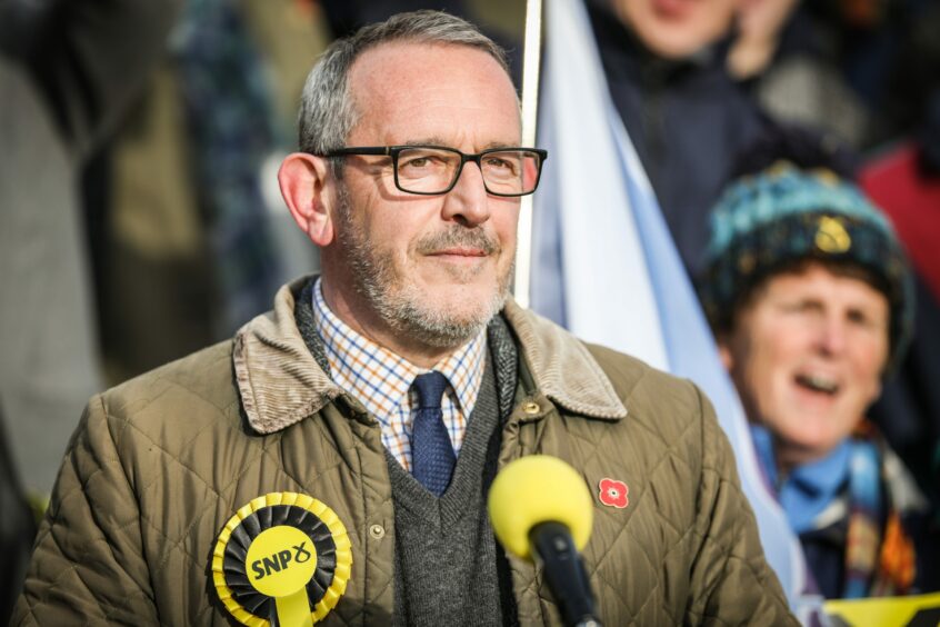 Dundee MP Stewart Hosie standing behind a microphone, wearing SNP rosette, with SNP supporters in the background.