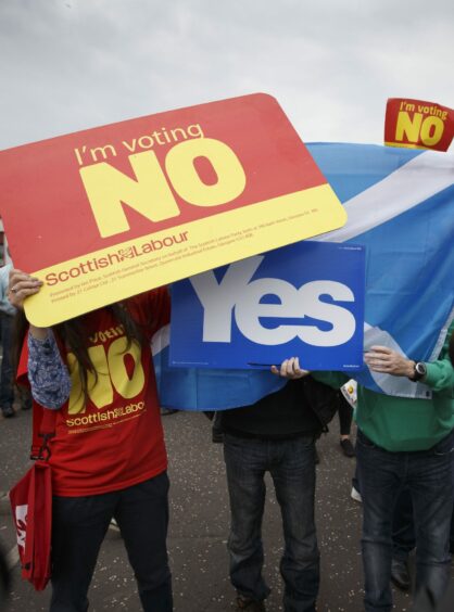 photo shows campaigners during the 2014 Scottish independence referendum. A person in a red Scottish Labour T shirt is holding a placard which reads 'I'm voting NO' in front of two people holding a Saltire flag and a placard with the word 'Yes'.