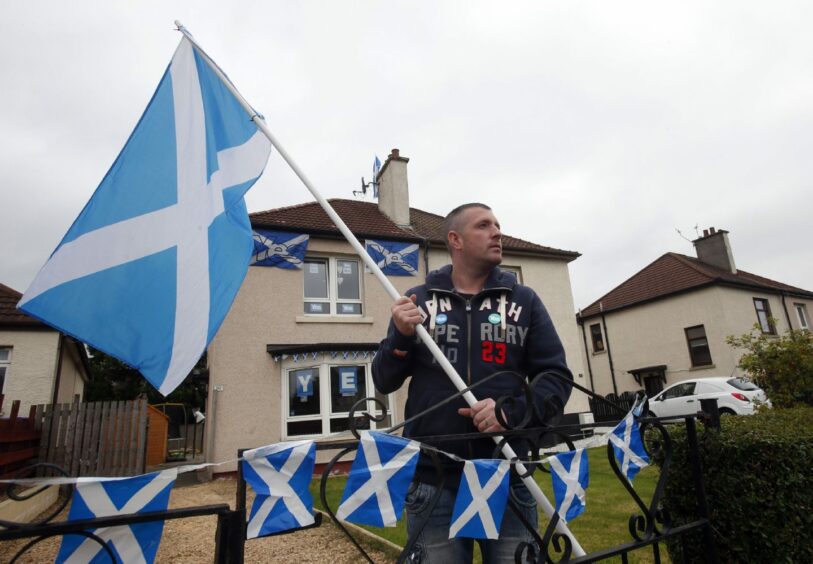 Photo shows a man outside his house waving a Saltire flag, with Saltire bunting on his fence and Saltires and Yes stickers in all of the windows.