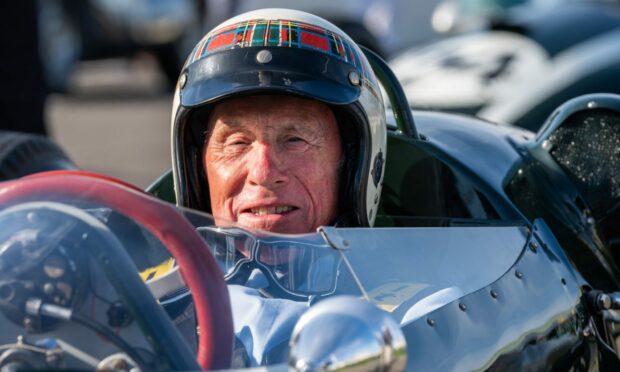 Sir Jackie Stewart, wearing his trademark tartan and white helmet, sits behind the red steering wheel of a classic race car at the annual vintage Revival event at Goodwood.