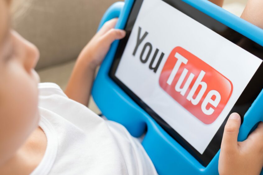 photo shows a young child holding a tablet with the YouTube logo on the screen.