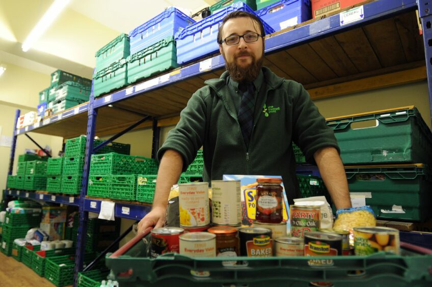 photo shows Ewan Gyrr, founder of Dundee foodbank, in front of shelves containing crates of food items.