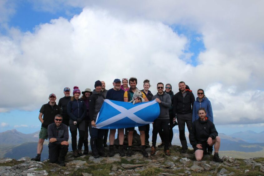 Ross Cunningham with a Saltire flag and a large group of friends on a mountain top.