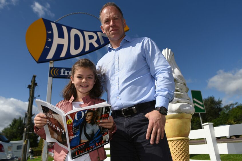 Kenny Farquharson with daughter Gabi, who is holding a copy of Vogue. Gabi is much younger in this photo. They are standing beside a roadside sign for The Horn.