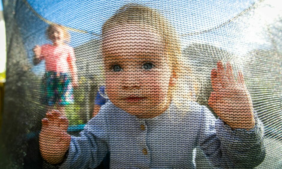 A almost 2-year-old girl with blue eyes and blonde hair looking through the safety net of a trampoline