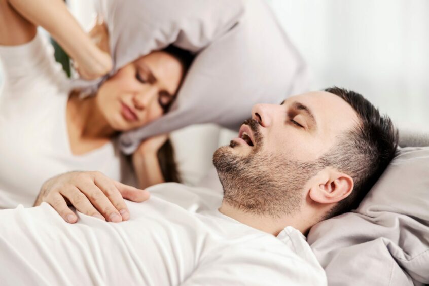 a man snoring in bed whilst a woman covers her ears with a pillow
