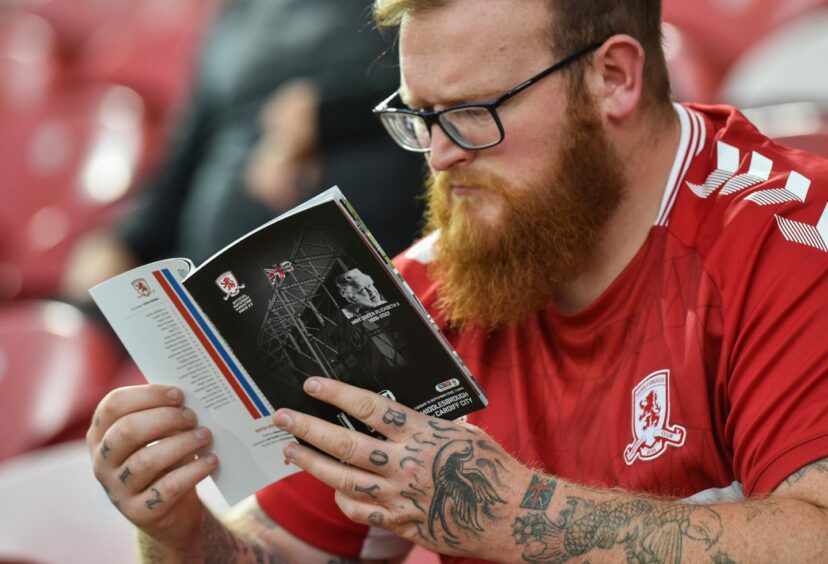 Photo shows a football fan in a red Middlesbrough top reading a matchday programme with an image of the Queen on the cover.