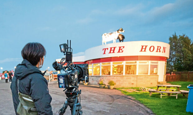 person standing behind film camera outside The Horn milk bar with model cow on roof