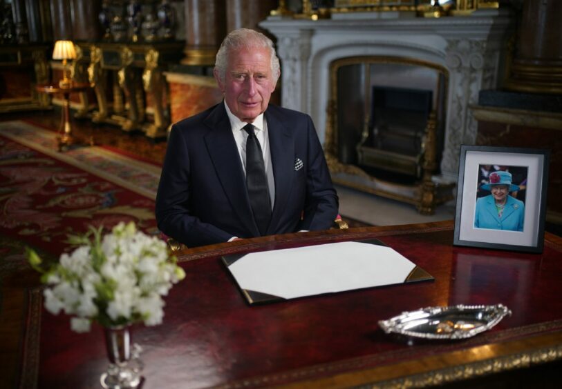 Photo shows King Charles seated at a desk in Buckingham Palace, with a photo of his late mother, the Queen., beside him.