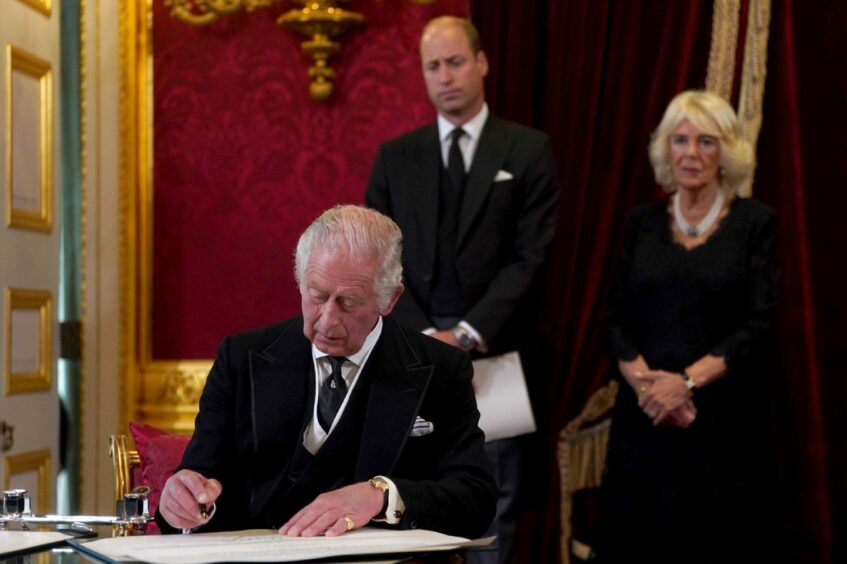 Photo shows King Charles at a table signing an oath, watched by his son Prince William and his wife Camilla.