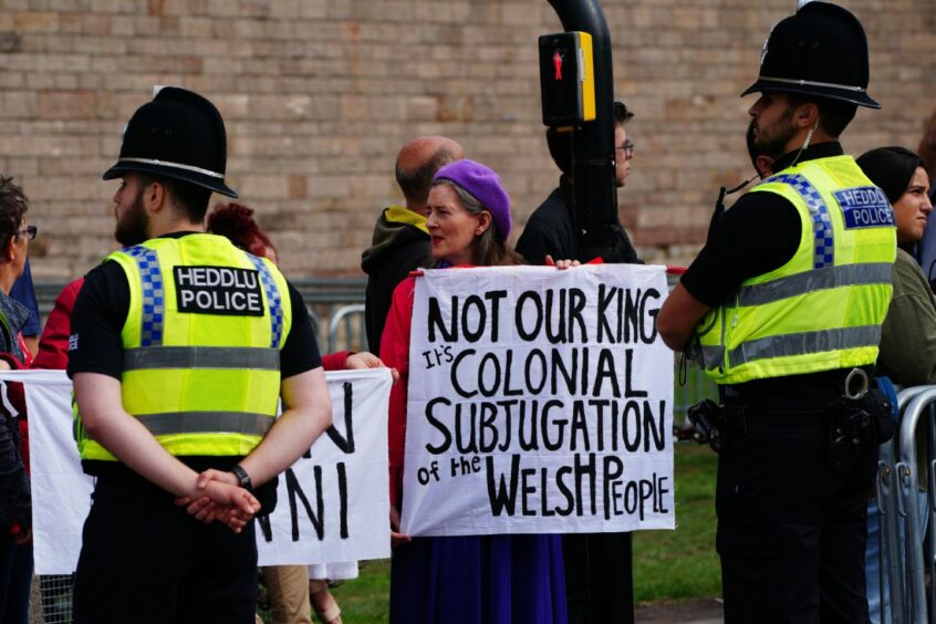 photo shows a woman holding a banner saying 'Not our king - it's colonial subjugation of the Welsh people' among a crowd of people while two police officers look on.