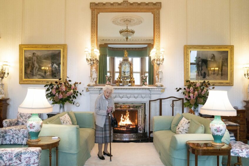 Photo shows the queen with a walking stick in front of a coal fire in the drawing room at Balmoral Castle.