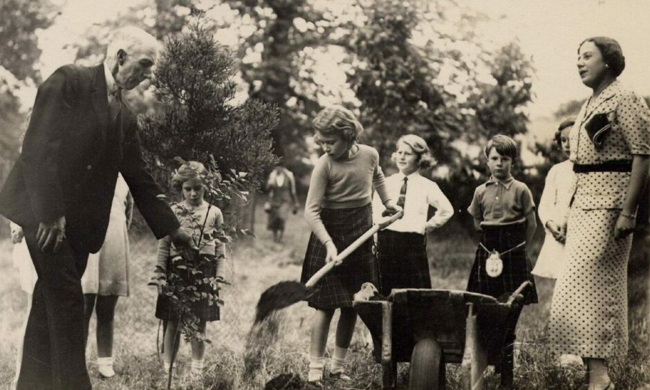 black and white photo shows the queen, as a young girl, planting a tree at Glamis, surrounded by five children and two adults.
