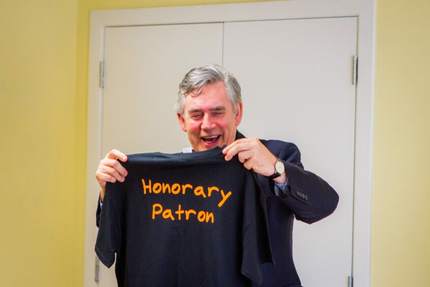 photo shows a smiling Gordon Brown holding a T shirt with the words 'Honorary Patron'.