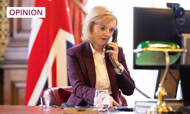 Photo shows Liz Truss holding a telephone receiver, while seated at a desk with a Union Jack hanging behind her.