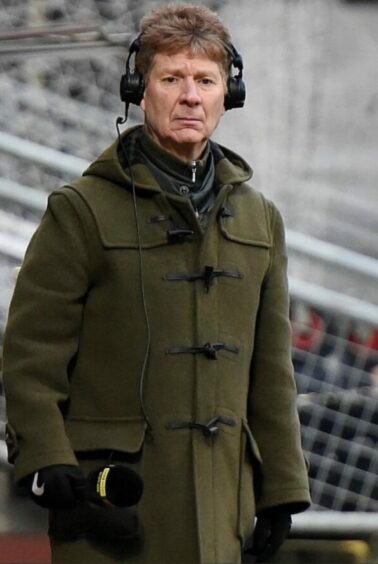 Jim Spence standing on the touchline at a football match during his days as a football commentator. He is wearing an olive green duffel coat, with a set of headphones over his ears and a microphone in hand.