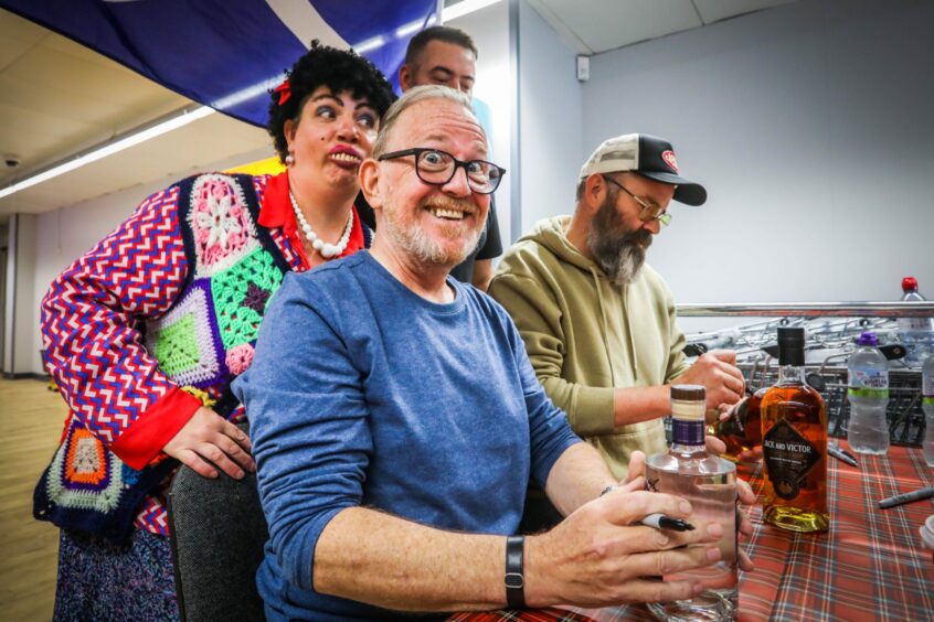 Photo shows Still Game stars Ford Kiernan and Greg Hemphill signing bottles of whisky and gin at a table, with a woman dressed as Edith, a character from the show, standing behind them.