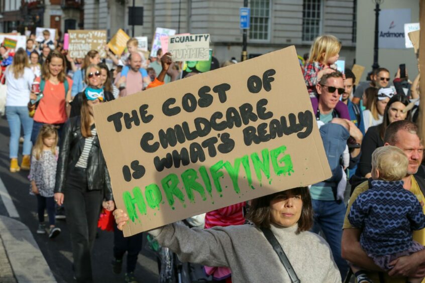 photo shows a large crowd of protesters carrying placards with slogans such as 'The cost of childcare is what's really horrifying'.