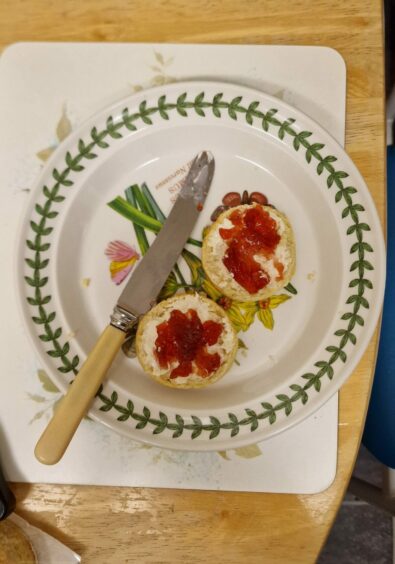 Photo shows a plate and knife, with a scone cut in half and spread with butter and jam.