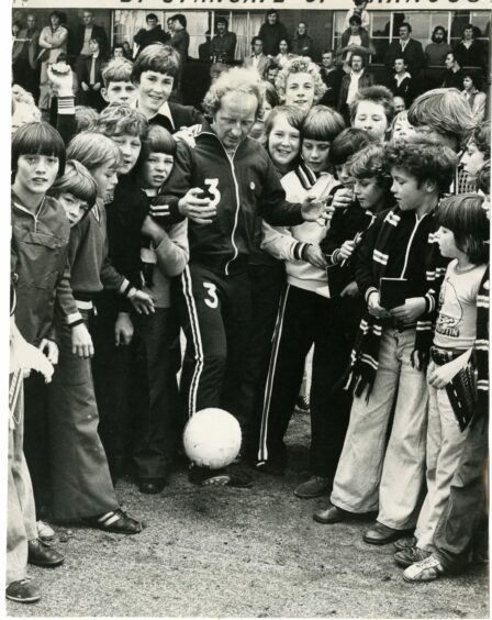 black and white photo shows Jimmy Johnstone playing with a football surrounded by children in 1970s fashions.