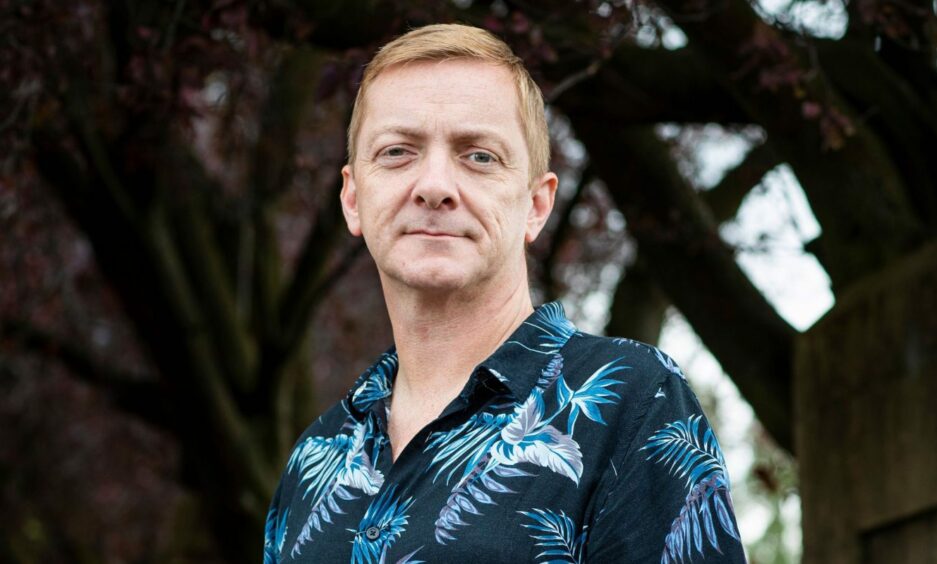 Image shows: author Doug Johnstone standing under a tree. He has red hair and is wearing a blue shirt with tropical, leafy print.