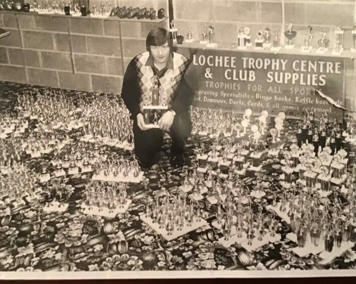 Black and white photo shows a man surrounded by hundreds of trophies and a sign saying Lochee Trophy Centre and Club Supplies.