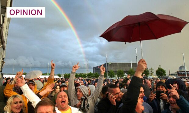 Photo shows a large crowd at an outdoors concert in Slessor Gardens, Dundee. One fan is holding an umbrella and there's a rainbow in the distance.