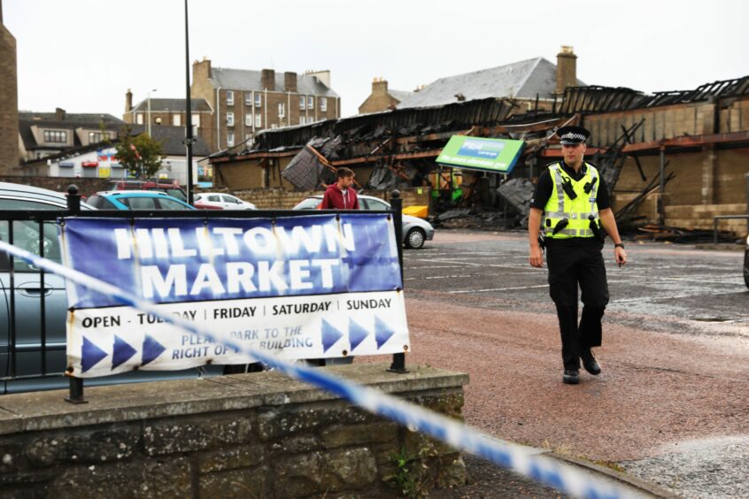 Photo shows a police officer walking past a sign for the Hilltown market, with the fire-damaged market buildings behind him.