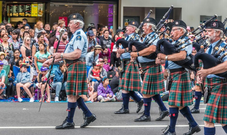 photo shows a pipe band parading through the streets of Aukland, New Zealand, watched by a large crowd of people.