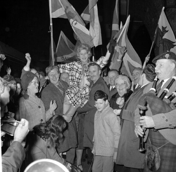 black and white photo shows Winnie Ewing being carried on the shoulders of two men in overcoats, surrounded by flag-waving supporters and a bagpiper.