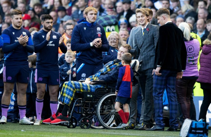 photo shows Doddie Weir in a wheelchair with Scotland rugby players applauding in the background.