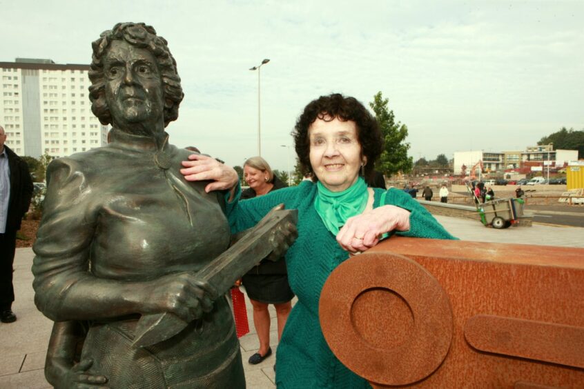 Photo shows Stella Carrington next to a bronze statue of a mill worker with the Lochee multi-storey flats behind her.