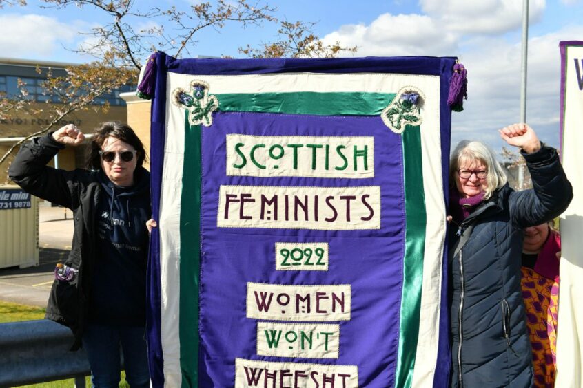 Photo shows two women with raised fists next to a large embroidered banner with the words 'Scottish feminists 2022: women won't wheesht'.