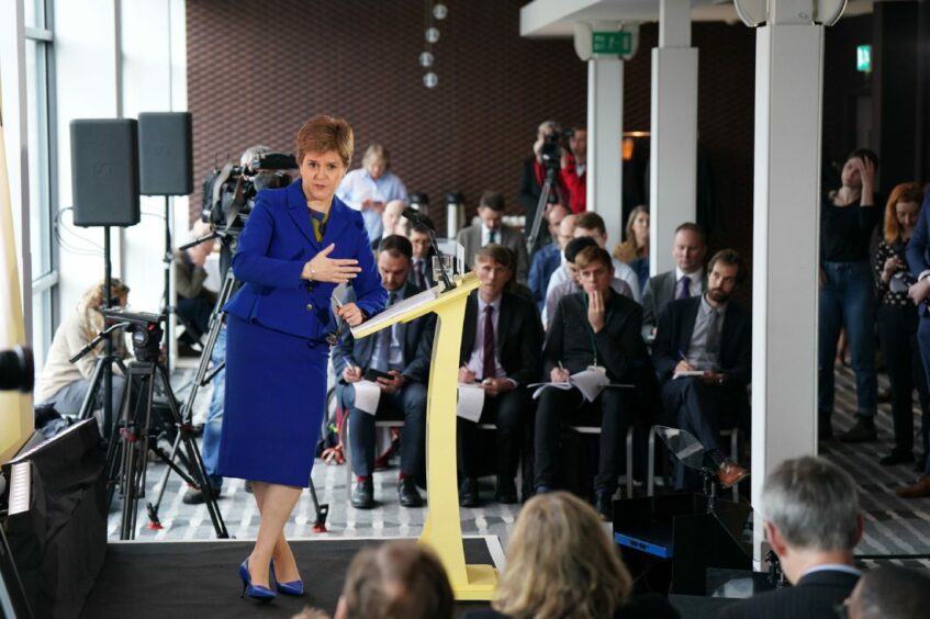 Nicola Sturgeon in blue suit standing at a lectern in front of a room full of reporters at a press conference in an Edinburgh hotel.