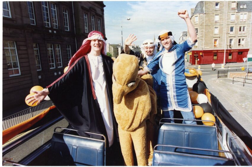 photo shows three men in desert robes and flowing headgear on an open-topped bus with someone in a camel costume.