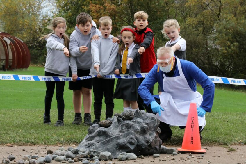 Photo shows children behind police tape pointing at a large lumpy rock, while a man in safety glasses and apron kneels down to take a closer look.