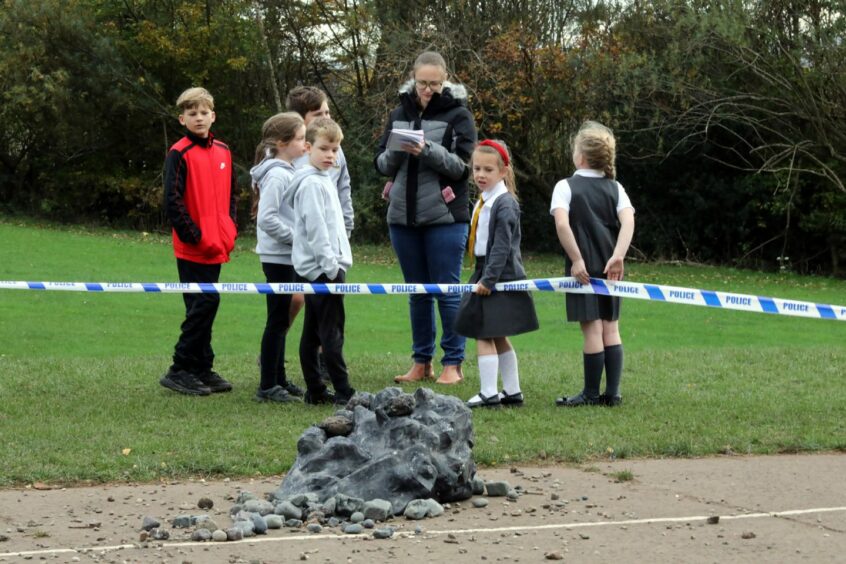 photo shows children behind police tape talking to a reporter with a notebook and looking at a lump of rock in the playground.