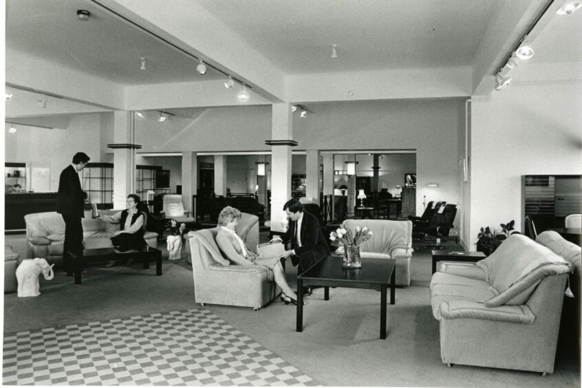 black and white photo shows suited shop assistants and smartly dressed women seated on a furniture display inside the store in 1987.