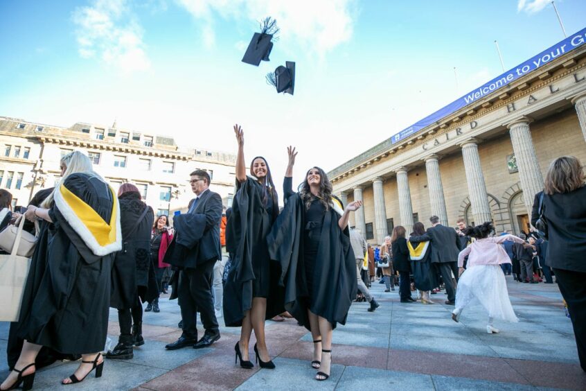 image shows two young women in gowns throwing their mortar boards in the air as part of a large group of graduating Dundee University students outside the Caird Hall.