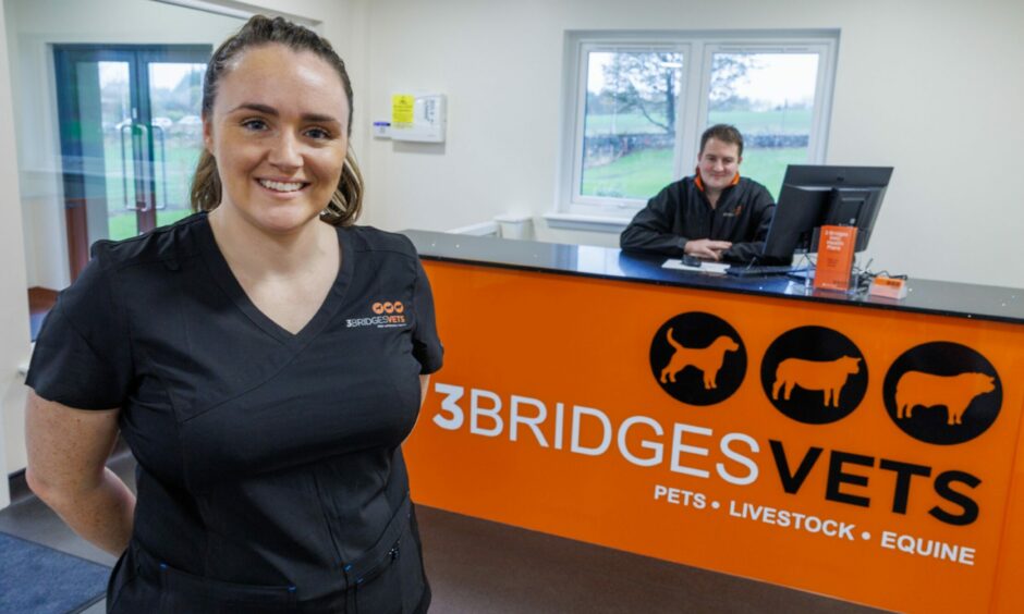 A woman in a vet uniform in front of an orange reception with the 3 Bridges Vets logo and a man behind the desk.