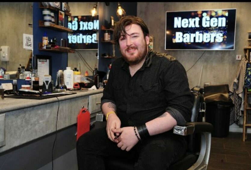 Photo shows Keiran McGarrity sitting in a barber's chair in front of a sign for Next Gen Barbers.