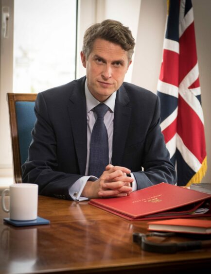 photo shows Gavin Williamson seated at a desk with a union jack flag behind him.