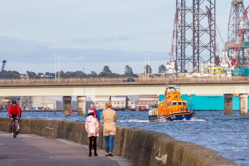 photo shows people walking and cycling beside the River Tay as a RNLI lifeboat passes under the Tay Bridge .