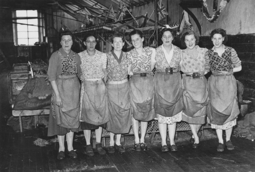 Black and white photo shows seven women in overalls, standing in front of mill machinery.