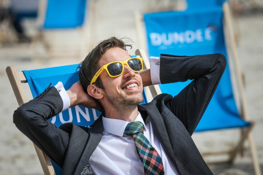 photo shows Dundee City Council leader John Alexander wearing yellow framed sunglasses and sitting on a blue deckchair with Dundee written on it.