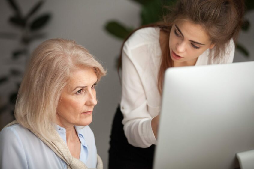 Photo shows an older woman looking at a computer screen, while a younger woman explains something to her.