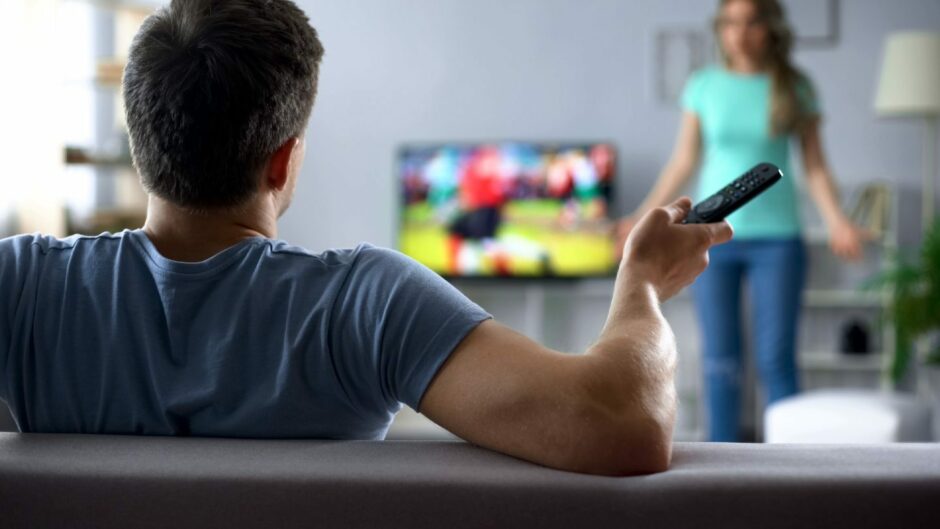 Photo shows the back of a man's head and shoulders as he watches football on TV while a woman waves her arms.